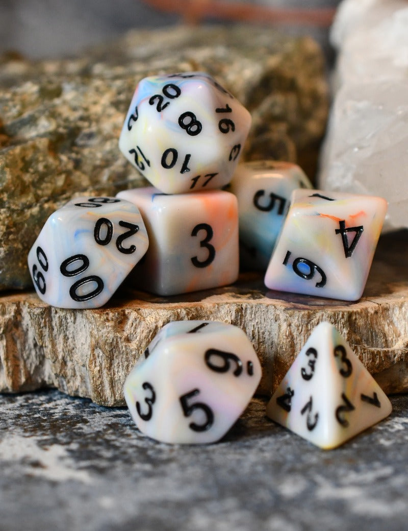 A set of marbled polyhedral dice with black printed numbers, arranged on a rustic wood slice with natural rocks in the background.