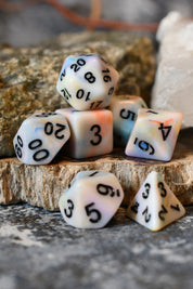 A set of marbled polyhedral dice with black printed numbers, arranged on a rustic wood slice with natural rocks in the background.
