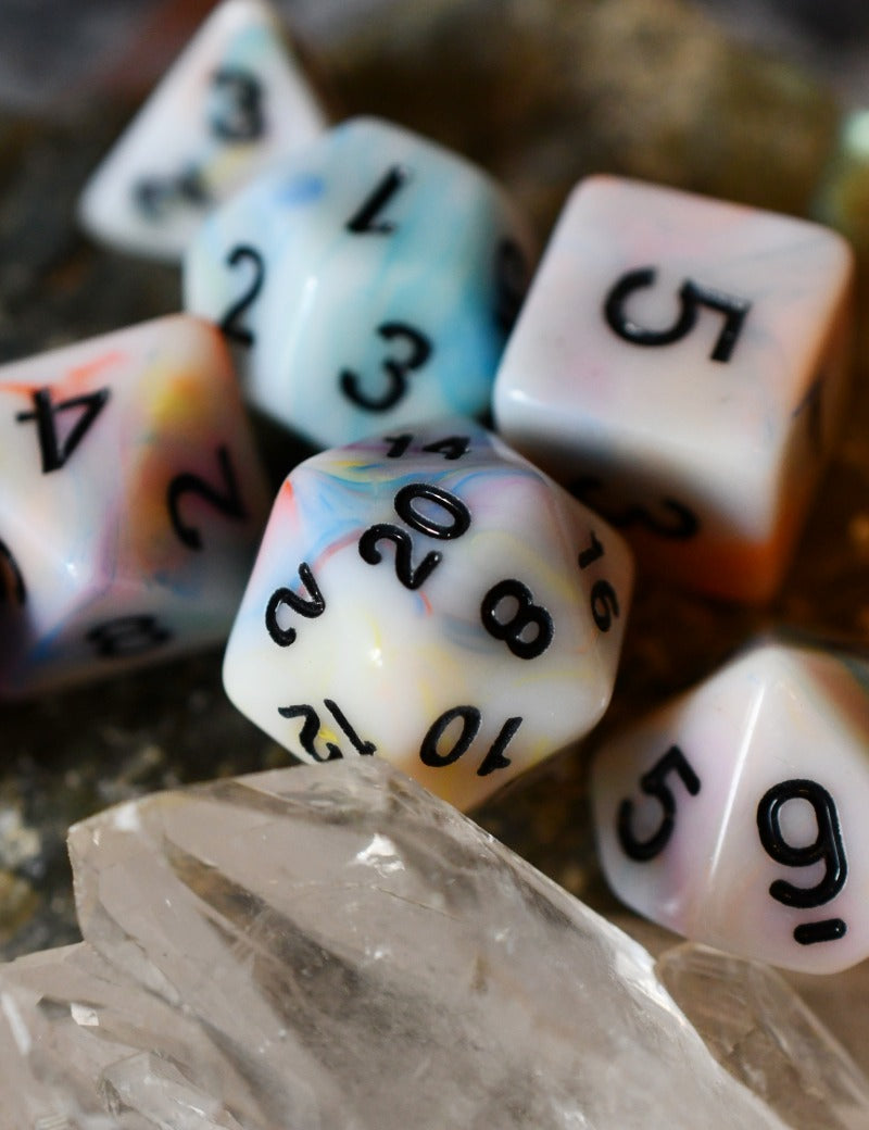 A set of marbled polyhedral dice with black printed numbers, arranged on a clear crystal cluster with a blurred natural stone background.