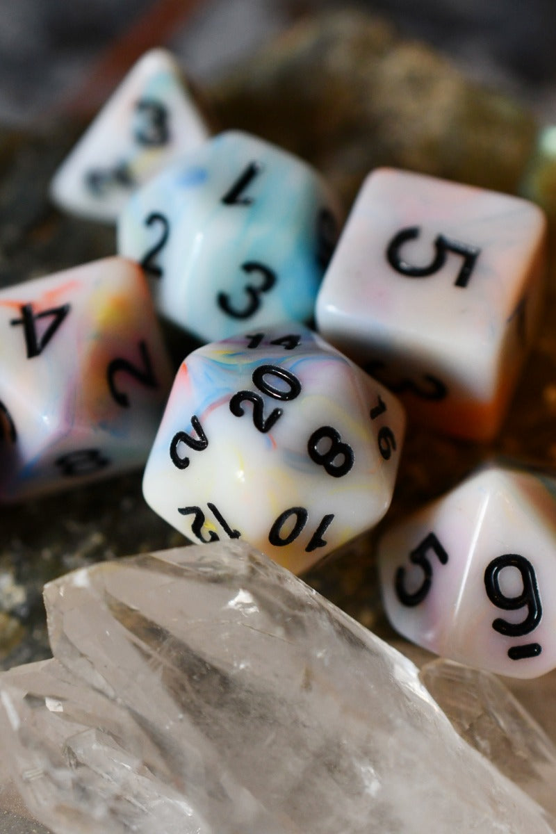 A set of marbled polyhedral dice with black printed numbers, arranged on a clear crystal cluster with a blurred natural stone background.