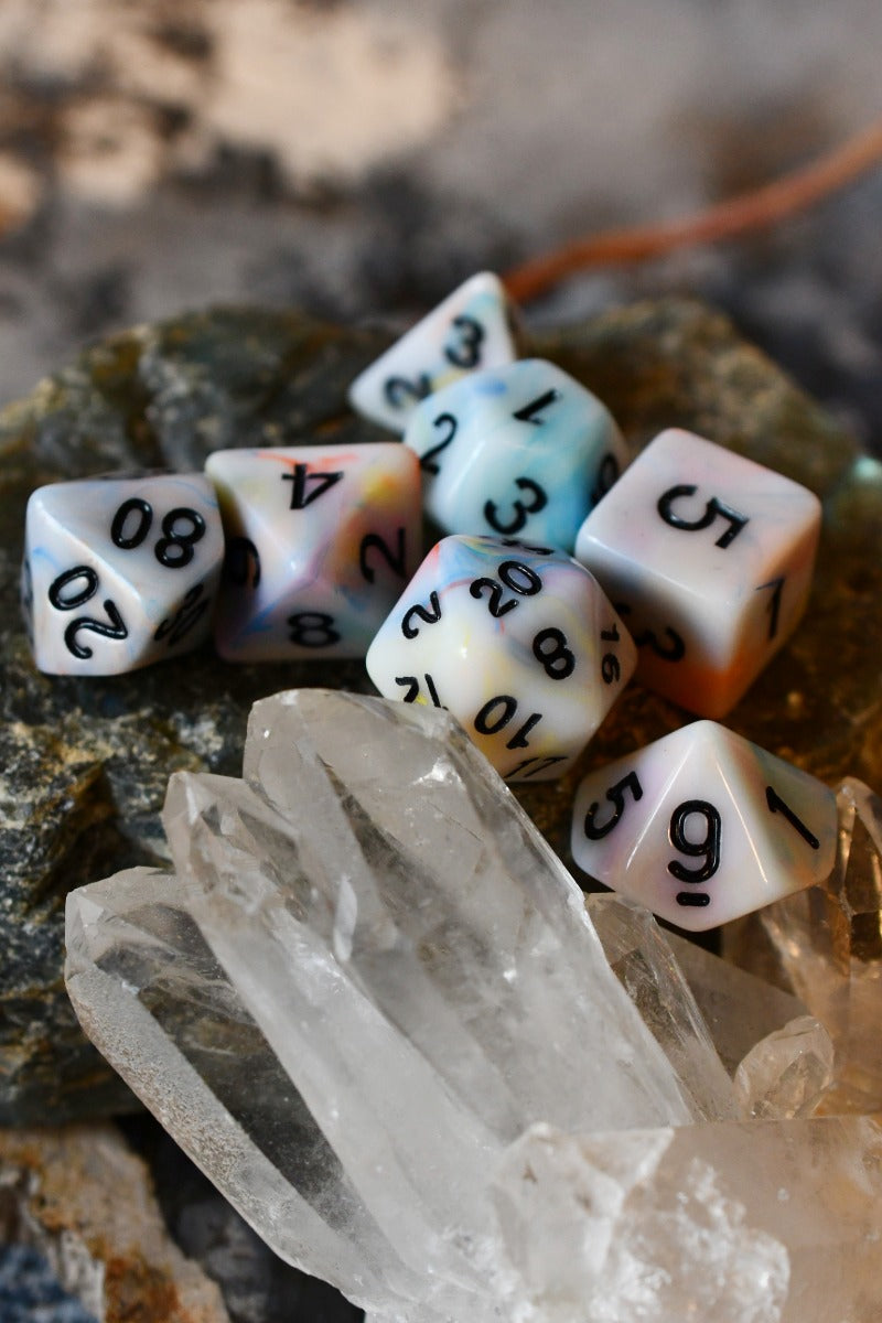 A set of translucent polyhedral gaming dice with black printed numbers, displayed on rocks and crystals. The dice feature a pastel gradient design shifting between white, light blue, pink, and orange.