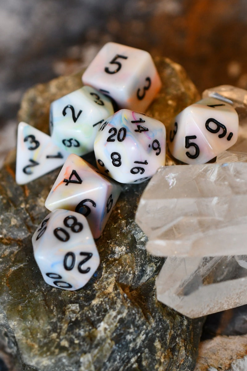 A set of white polyhedral gaming dice with black printed numbers, arranged on a textured rock next to a clear crystal point.