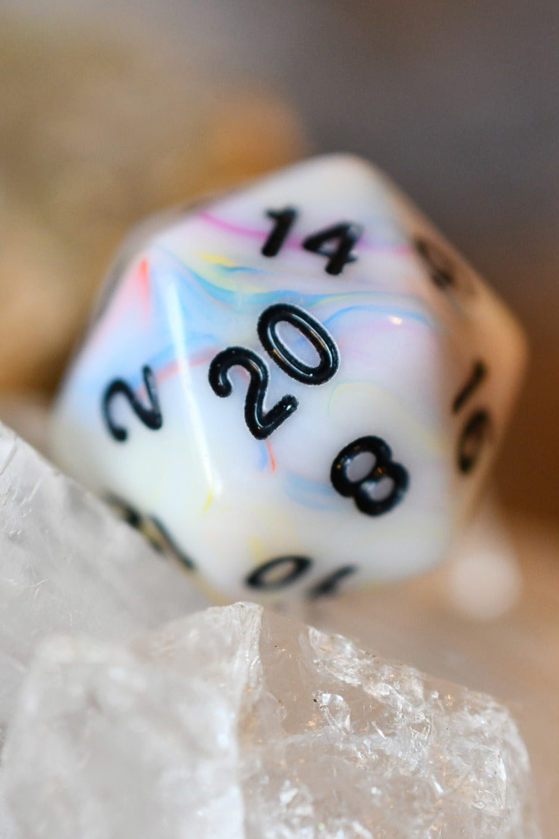 A close-up of a marbled polyhedral die, likely a d20, with black printed numbers and a white base featuring pastel swirls of pink, blue, and yellow. It is resting on clear quartz crystals against a soft-focus neutral background.