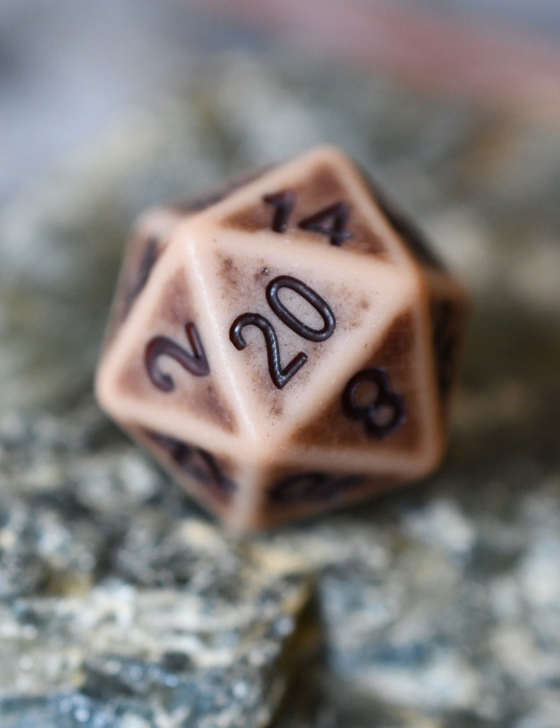 A 20-sided d20 polyhedral gaming die with a warm beige base and dark brown engraved numbering, resting on a textured gray stone surface.