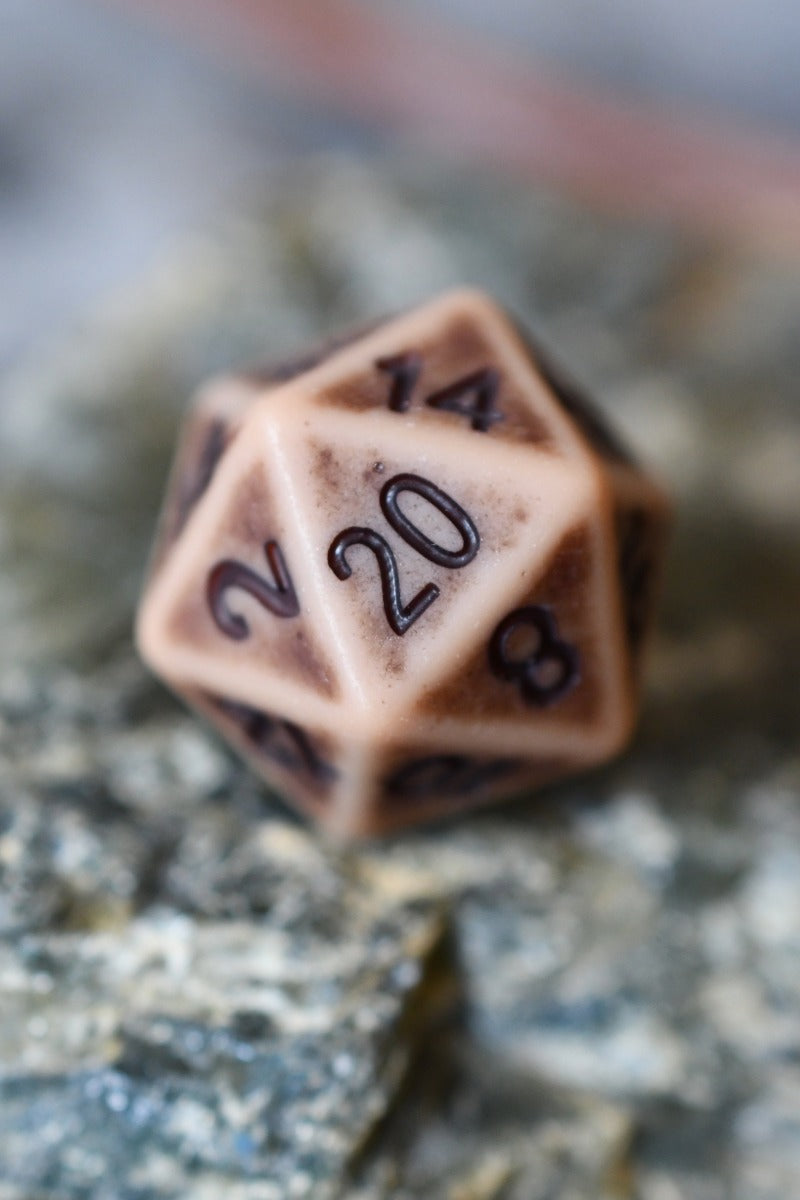 A 20-sided d20 polyhedral gaming die with a warm beige base and dark brown engraved numbering, resting on a textured gray stone surface.