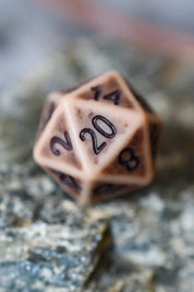 A 20-sided d20 polyhedral gaming die with a warm beige base and dark brown engraved numbering, resting on a textured gray stone surface.