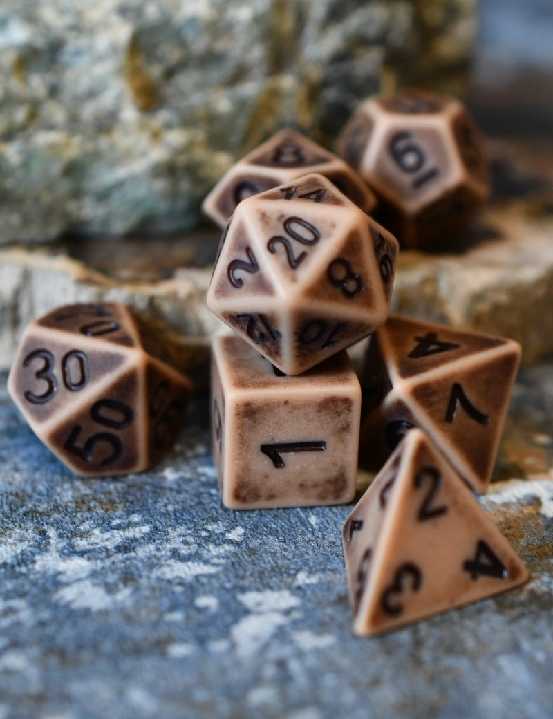 A set of brown polyhedral tabletop gaming dice with black printed numbers, arranged on a textured stone surface with a blurred natural rock background.