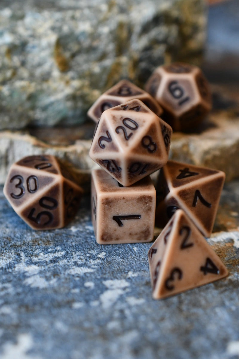 A set of brown polyhedral tabletop gaming dice with black printed numbers, arranged on a textured stone surface with a blurred natural rock background.