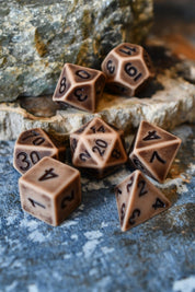 A set of brown polyhedral tabletop gaming dice with black printed numbers, arranged on a textured blue-gray surface with rocks in the background.