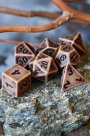 A set of tan polyhedral gaming dice with black printed numbers, arranged on a gray stone with blurred brown branches in the background.