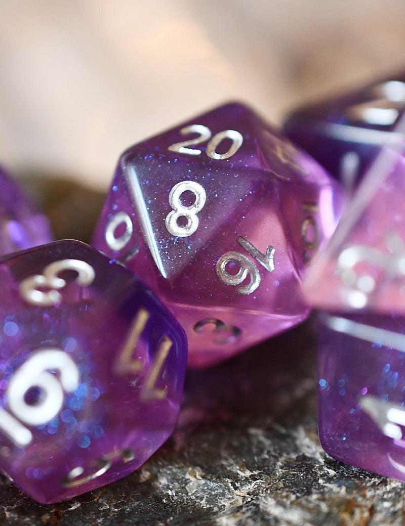 Purple glitter polyhedral gaming dice with silver numbers, including a d20, resting on a dark textured surface with a blurred warm background.