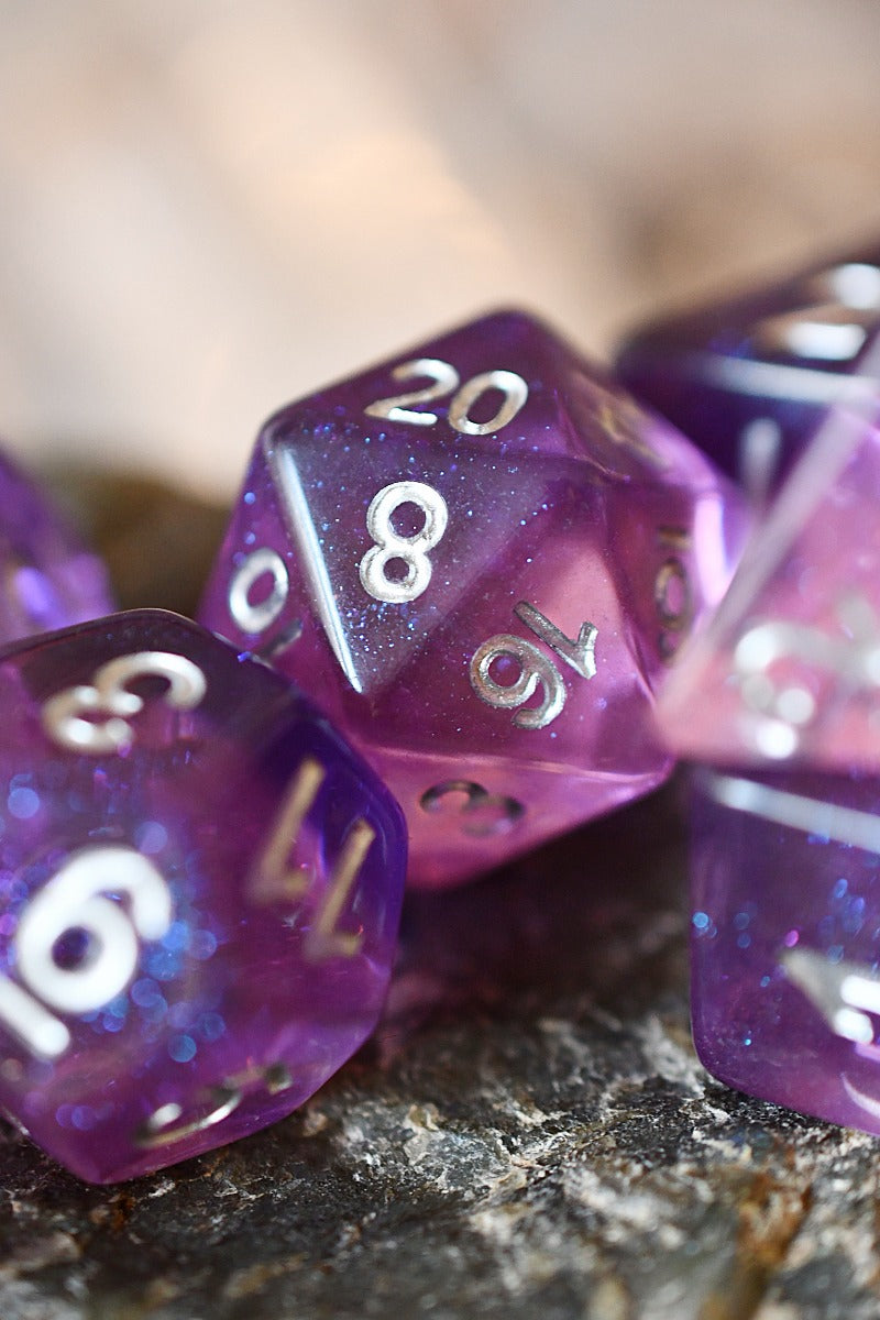 Purple glitter polyhedral gaming dice with silver numbers, including a d20, resting on a dark textured surface with a blurred warm background.