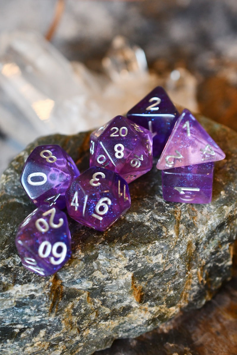 A set of translucent purple polyhedral gaming dice with white numbering, arranged on a textured gray rock with blurred natural background elements.