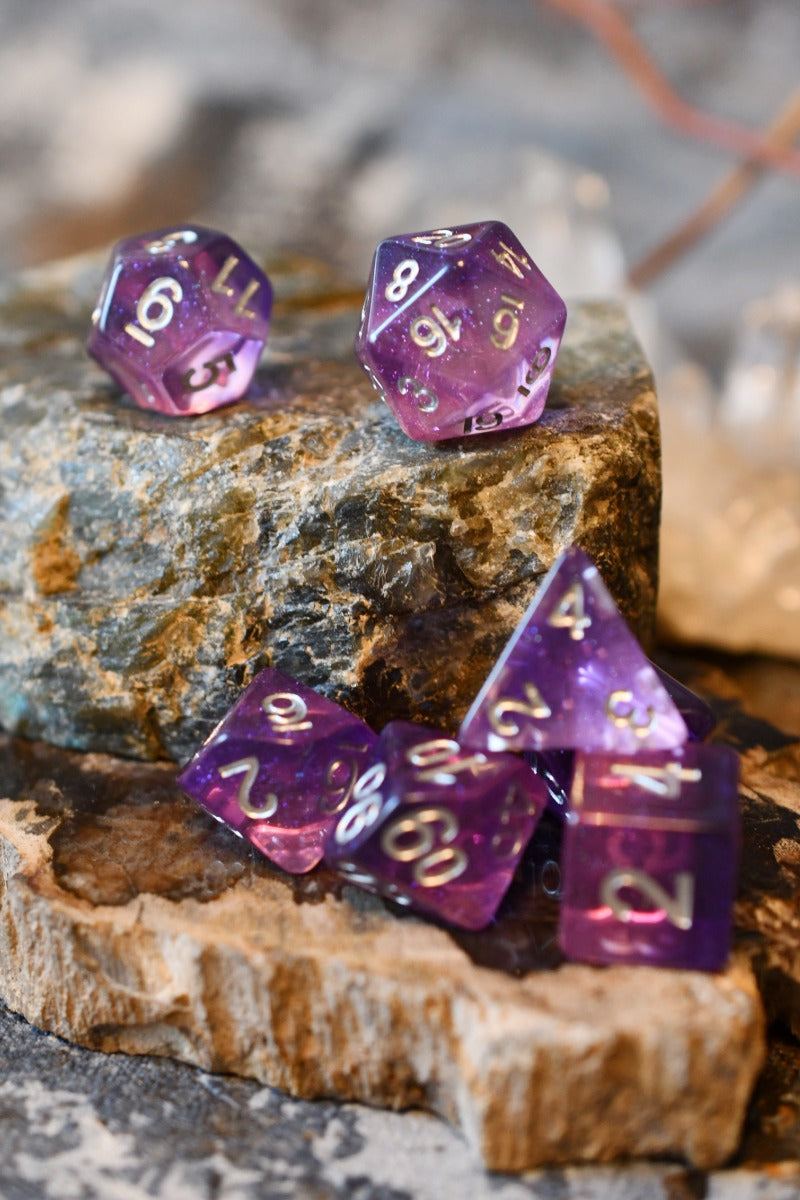 Purple translucent polyhedral tabletop gaming dice with silver numbers, arranged on stacked natural stones and wood slices.