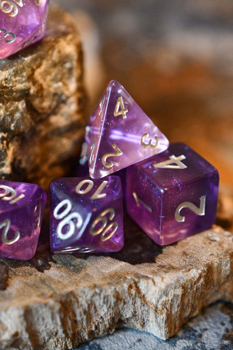 Purple translucent polyhedral gaming dice with metallic gold numbers, arranged on a rustic wood slice with a blurred natural background.