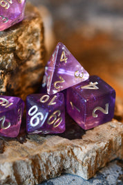 Purple translucent polyhedral gaming dice with metallic gold numbers, arranged on a rustic wood slice with a blurred natural background.