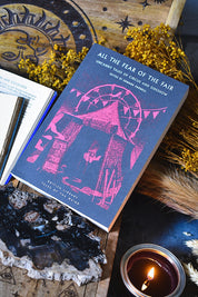 A book titled "All the Fear of the Fair" lies open on a rustic wooden surface surrounded by dried yellow flowers, a lit candle, crystals, and a printed celestial design. The visible cover is dark blue with pink line art and white text.
