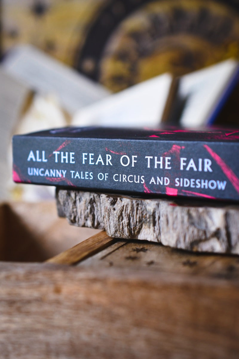 A close-up of a book titled 'All the Fear of the Fair: Uncanny Tales of Circus and Sideshow' resting on a rustic wood slice, with a blurred background.