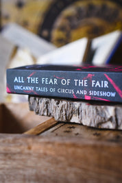 A close-up of a book titled 'All the Fear of the Fair: Uncanny Tales of Circus and Sideshow' resting on a rustic wood slice, with a blurred background.