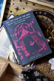 A hardcover book titled "All the Fear of the Fair" lies on a rustic wooden tray surrounded by occult-themed props including crystals, dried herbs, an old book, and a star-patterned coaster.