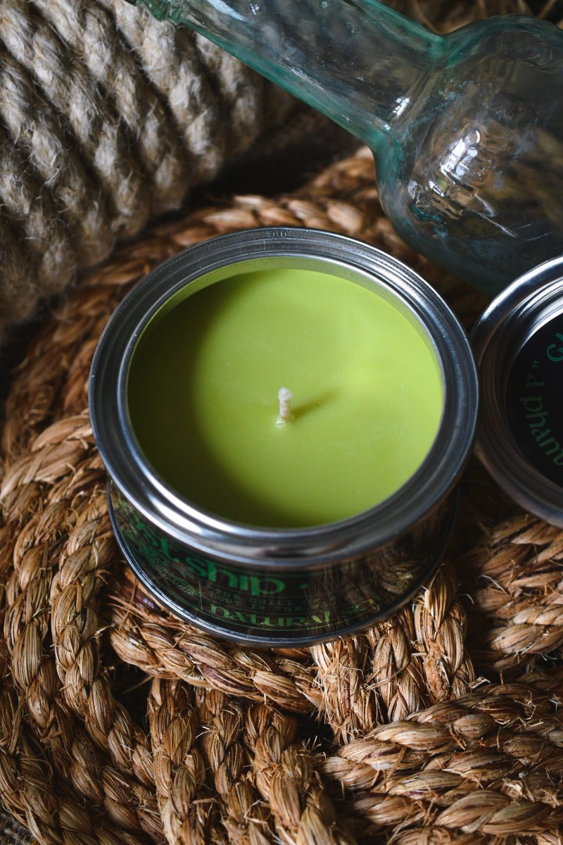 A green wax candle in a silver metal tin with a white wick, styled on a woven jute mat with a glass bottle in the background.