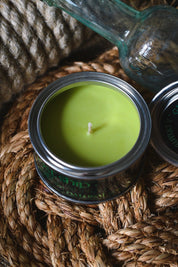 A green wax candle in a silver metal tin with a white wick, styled on a woven jute mat with a glass bottle in the background.
