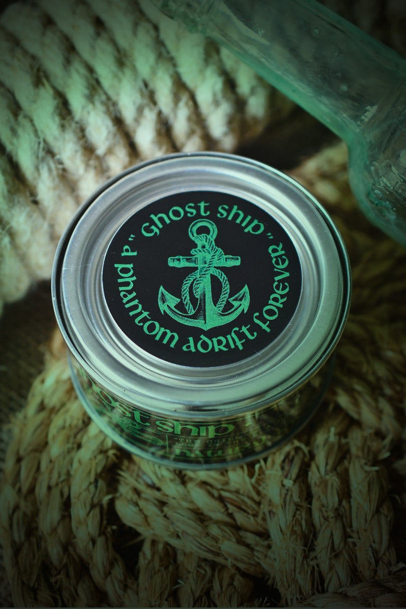 A small clear plastic jar with a silver lid sits on a bundle of natural rope. The lid has a black label with a green anchor graphic and the words 'Ghost Ship' and 'phantom dorft forever'.