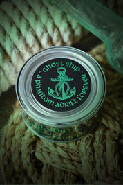 A small clear plastic jar with a silver lid sits on a bundle of natural rope. The lid has a black label with a green anchor graphic and the words 'Ghost Ship' and 'phantom dorft forever'.