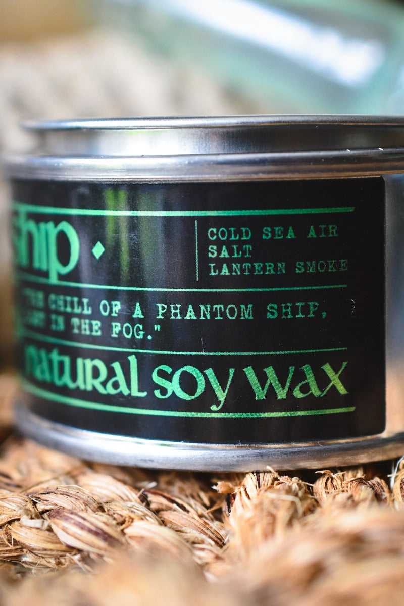 A close-up of a cylindrical tin candle labeled 'natural soy wax' with a black label and green text, resting on a bed of dried brown plant fibers. The label reads 'Ship' and 'Cold Sea Air Salt Lantern Smoke'.