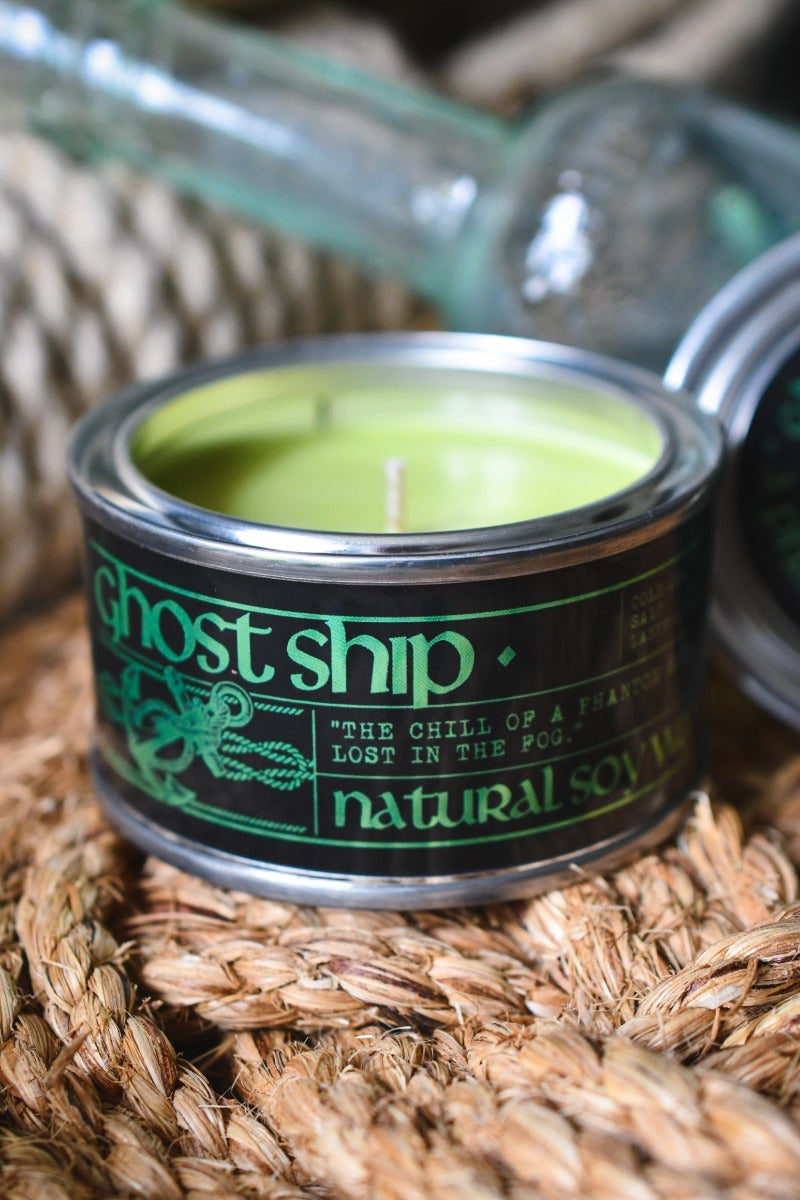 A small candle in a metal tin with a black label reading 'ghost ship' and 'natural soy wax,' filled with light green wax, set on a woven straw surface with glass bottles blurred in the background.