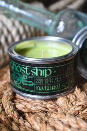 A small candle in a metal tin with a black label reading 'ghost ship' and 'natural soy wax,' filled with light green wax, set on a woven straw surface with glass bottles blurred in the background.