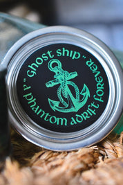 A close-up of a round metal tin lid featuring a black background with a green anchor graphic and the words "Ghost ship" and "phantom drift forever" in green text, resting on a bed of brown shredded paper.