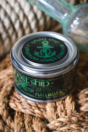 A small metal tin of Ghost Ship Natural Soap sits on a woven rope surface with a blurred glass bottle in the background. The tin has a silver lid with a black label featuring green text and an anchor graphic.