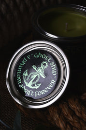 A close-up of a metal tin lid with a black label featuring a green anchor and the words 'Mourned F" Ghost Ship" drift forever,' with part of the candle jar and green wax visible in the background.