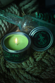 A lit green candle in a metal tin sits on coiled rope next to its lid, with a clear glass bottle in the background. The lid shows the brand name 'ghost ship' with an anchor graphic.