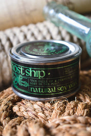 A small round tin of Ghost Ship Natural Soy Wax sits on a woven straw mat, with a blurred glass bottle in the background.