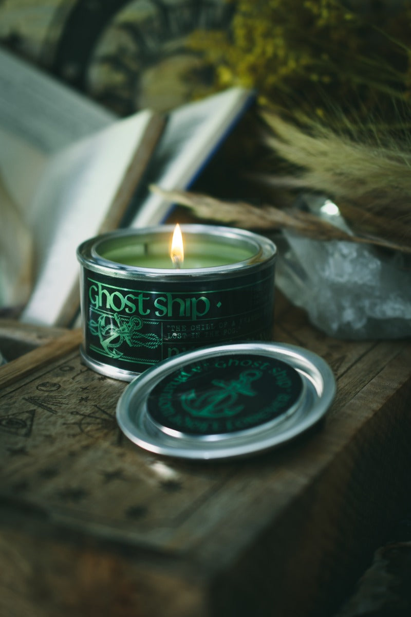 A lit green candle in a dark metal tin with the brand name Ghost Ship visible on the label, placed on a rustic wooden surface with its lid set beside it. The scene includes dried grasses and an open book in a moody, atmospheric setting.