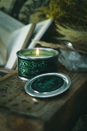 A lit green candle in a dark metal tin with the brand name Ghost Ship visible on the label, placed on a rustic wooden surface with its lid set beside it. The scene includes dried grasses and an open book in a moody, atmospheric setting.
