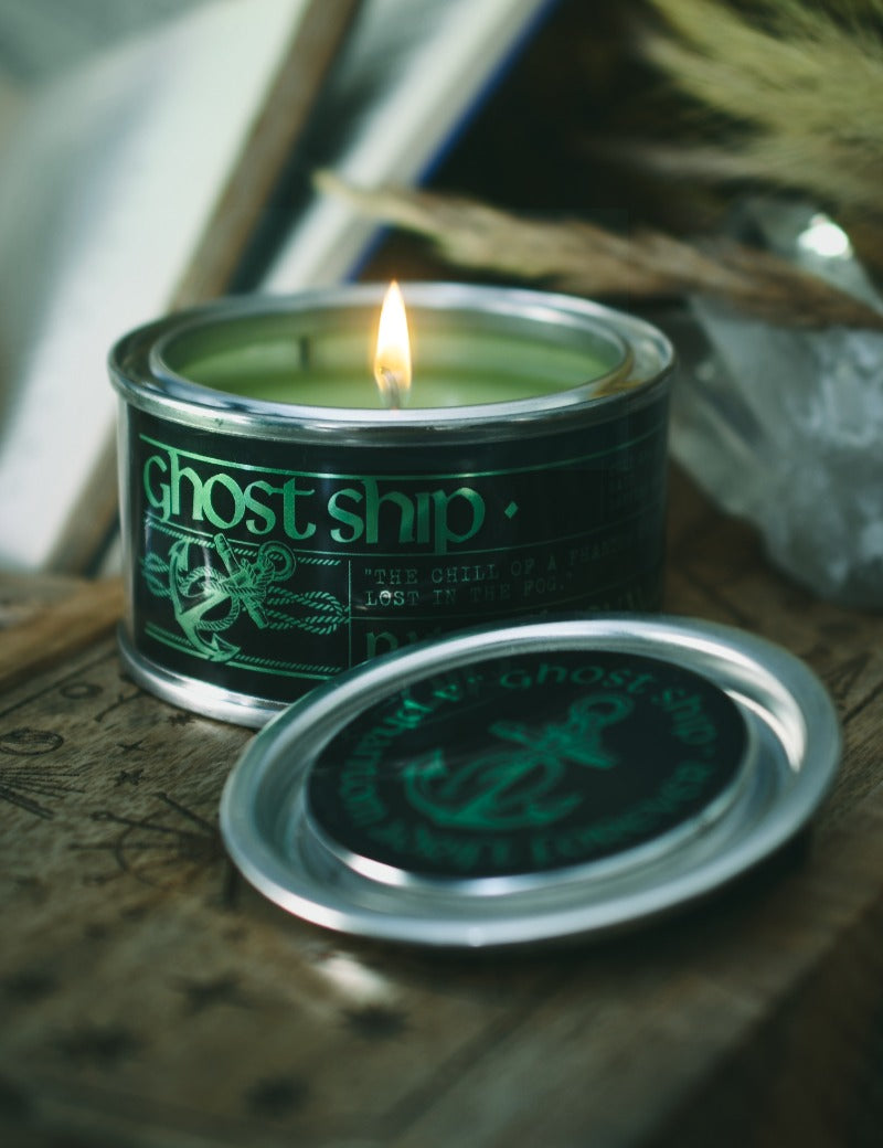 A lit candle in a silver tin with a dark green label reading 'ghost ship', placed on a rustic wooden surface with the matching lid in front. The background is softly blurred with dried grass and neutral tones.