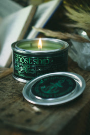 A lit candle in a silver tin with a dark green label reading 'ghost ship', placed on a rustic wooden surface with the matching lid in front. The background is softly blurred with dried grass and neutral tones.