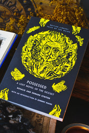 The book 'Possessed: A Lost Novel of the Occult' by Rosalie and Edward Synton, featuring a dark navy blue cover with a large yellow illustration of a possessed face and butterflies, styled on a rustic wooden surface.