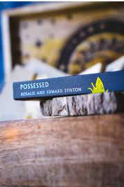 A close-up of the book cover of 'Posessed' by Rosalie and Edward Synton, resting on a rustic wooden slice with a blurred background.